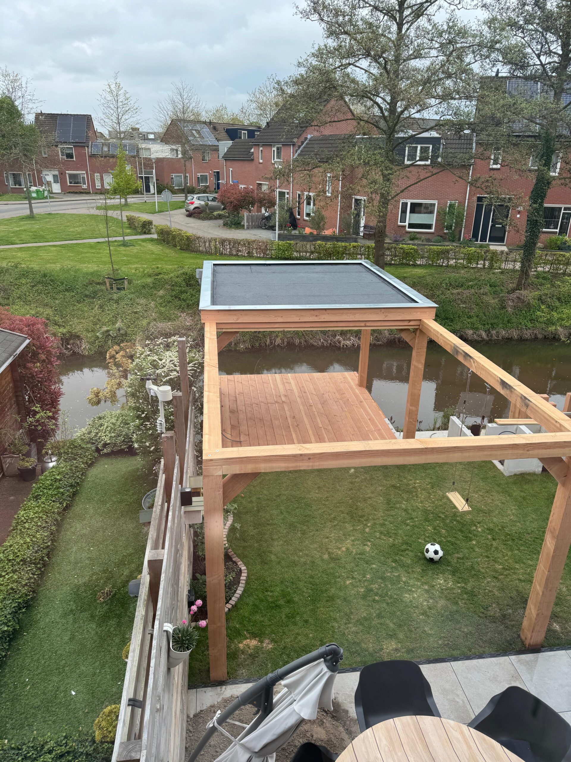 Vrijstaande douglas houten overkapping met plat dak en schommel in tuin aan het water, van bovenaf gezien